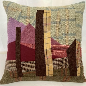 May include: Square decorative pillow with a patchwork design. The pillow features various textured fabrics in shades of green, brown, purple, and red. The design includes geometric shapes and patterns, creating a unique and artistic look. The pillow is likely for home decor.