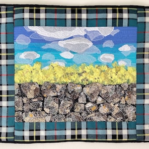 May include: A framed textile artwork featuring a landscape with a blue sky, white clouds, yellow grass, and a stone wall. The frame is a green, white, and black tartan.