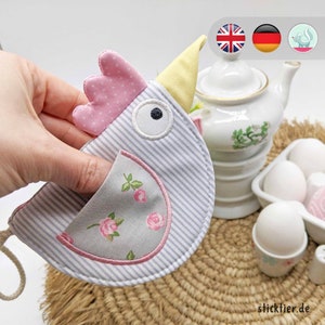 May include: A blue and white striped fabric chicken with a pink and white polka dot crest and a yellow beak. The chicken has a pocket on its chest with a floral pattern. The chicken is holding an egg in its pocket.