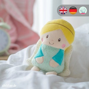 May include: A plush angel doll with blonde hair, a light blue dress with gold glitter, and white wings. The doll is sitting on a white bed.