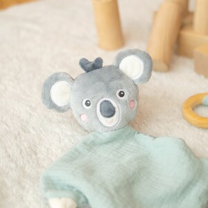 May include: A gray koala plush toy with a light blue muslin blanket. The koala has white ears, pink cheeks, and a dark gray nose. A wooden ring teether with a light blue fabric bunny ear design is also present.