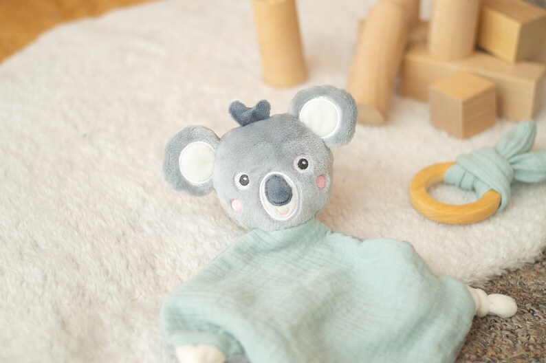 May include: A gray koala plush toy with a light blue muslin blanket. The koala has white ears, pink cheeks, and a dark gray nose. A wooden ring teether with a light blue fabric bunny ear design is also present.