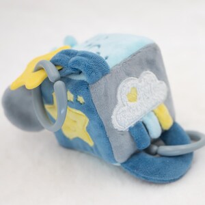 May include: A soft, plush baby toy cube in shades of blue, gray, and yellow. The cube features embroidered designs of a cloud, rainbow, and stars. It has plastic rings for easy grasping and play.