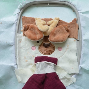 May include: An embroidery hoop holds a partially completed plush dog face. The dog has brown ears, a tan nose, and pink cheeks. The dog's body is white with brown spots. A maroon fabric is attached below the dog's face.
