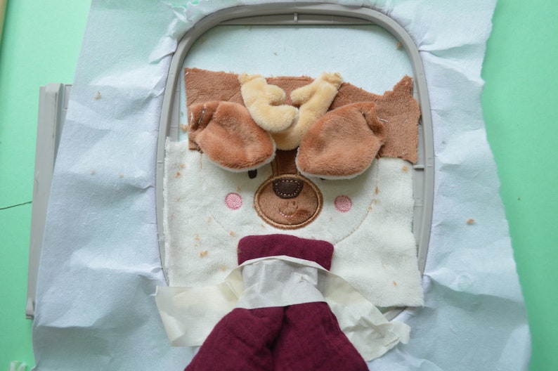 May include: An embroidery hoop holds a partially completed plush dog face. The dog has brown ears, a tan nose, and pink cheeks. The dog's body is white with brown spots. A maroon fabric is attached below the dog's face.