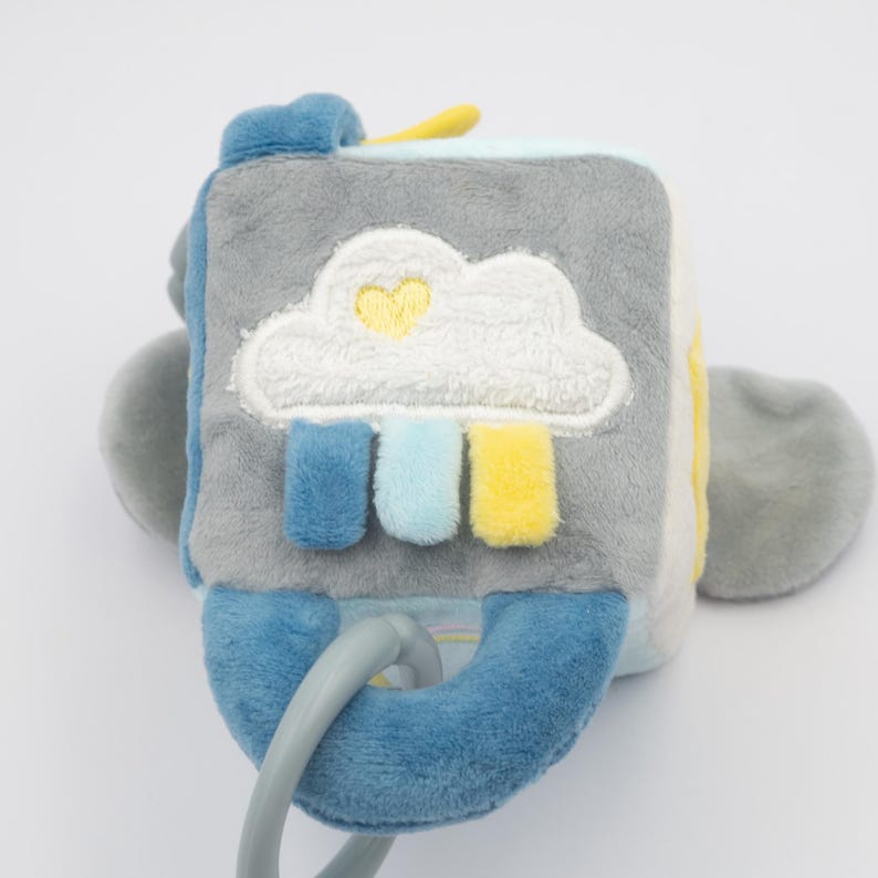 May include: A soft, plush baby toy in the shape of a cube. The cube is primarily gray with blue and yellow accents. A white cloud with a yellow heart is embroidered on one side, with blue, light blue, and yellow rectangles below. A blue loop is attached.