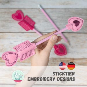 Könnte beinhalten: Rosa Filzpfeil-Stiftaufsätze mit herzförmigen Details. Die Pfeilspitzen und Federn sind mit dunkelrosa Nähten verziert. Das Bild enthält auch einen Bleistift und den Text "STICKTIER EMBROIDERY DESIGNS".