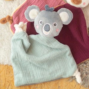 May include: A soft, gray koala plush toy with a light blue muslin blanket. The koala has embroidered facial features and pink cheeks. A burgundy and pink muslin blanket is visible in the background. The toy is designed for babies.