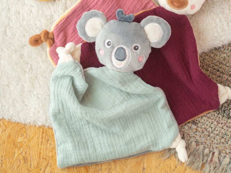 May include: A soft, gray koala plush toy with a light blue muslin blanket. The koala has embroidered facial features and pink cheeks. A burgundy and pink muslin blanket is visible in the background. The toy is designed for babies.