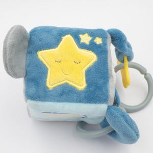 May include: A soft, blue baby toy cube with a yellow star and two smaller stars embroidered on one side. The star has a smiling face with closed eyes. The cube has a gray handle and a gray and yellow ring attachment.