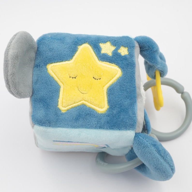May include: A soft, blue baby toy cube with a yellow star and two smaller stars embroidered on one side. The star has a smiling face with closed eyes. The cube has a gray handle and a gray and yellow ring attachment.
