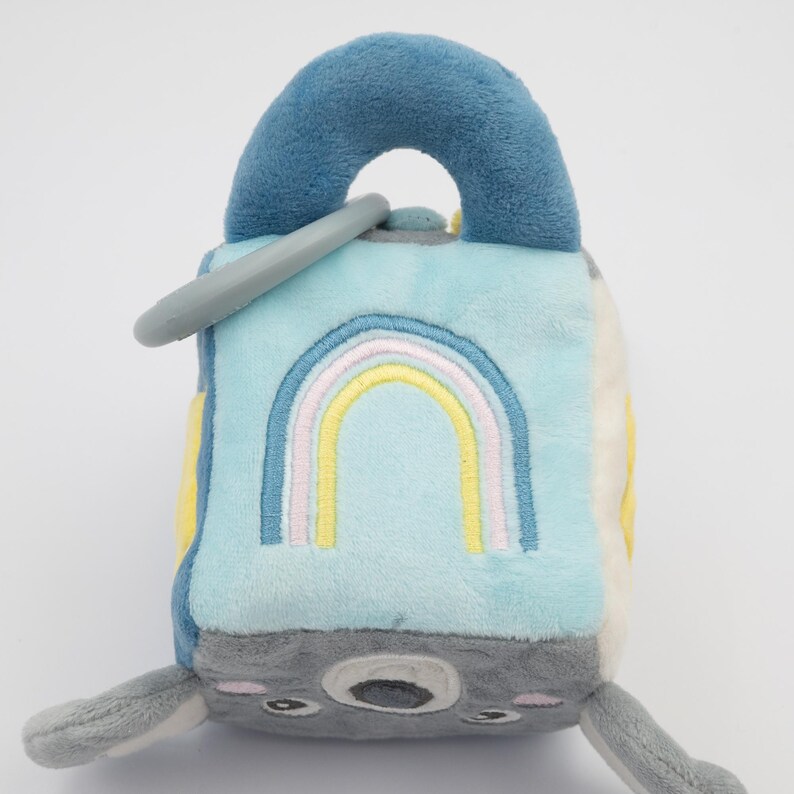 May include: A soft, plush baby toy in the shape of a cube with a blue handle and a gray ring. The cube features a light blue side with a rainbow design, gray and white sides, and a gray koala face.