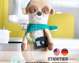 May include: A plush meerkat toy with a teal scarf and a camera. The meerkat has brown eyes and ears, a white face, and a small tuft of brown hair. The scarf has the word "NIO" embroidered on it. The toy is part of "STICKTIER EMBROIDERY DESIGNS".