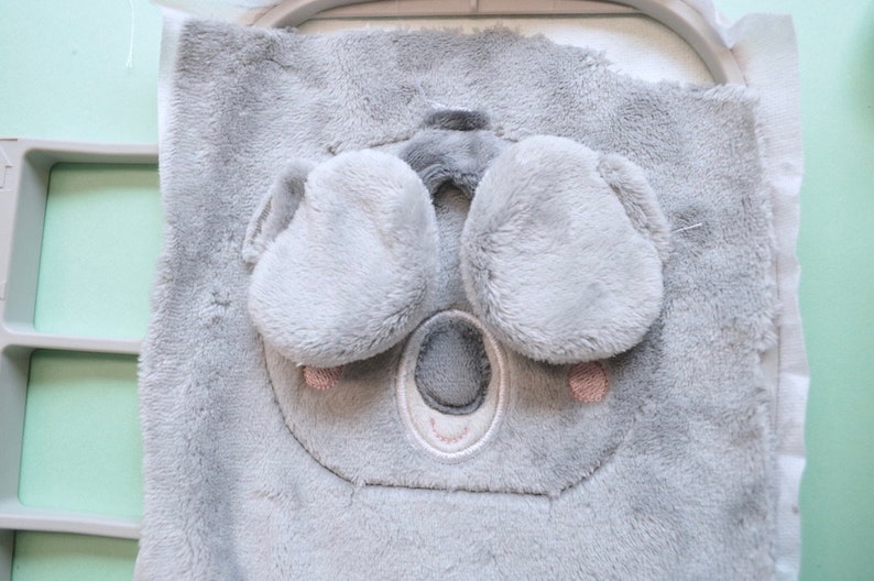 May include: A gray plush koala face with embroidered details, including a nose, eyes, and ears. The koala is part of a larger fabric piece, possibly for a baby item. The design is in an embroidery hoop.