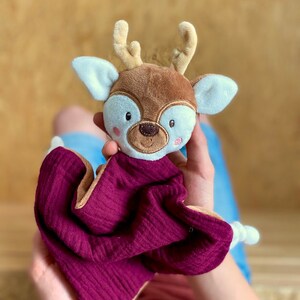 May include: A baby comforter in the shape of a deer. The deer's head is brown with beige antlers and white-lined ears. The comforter is a deep burgundy with a beige border. This soft toy is designed for infants and young children.