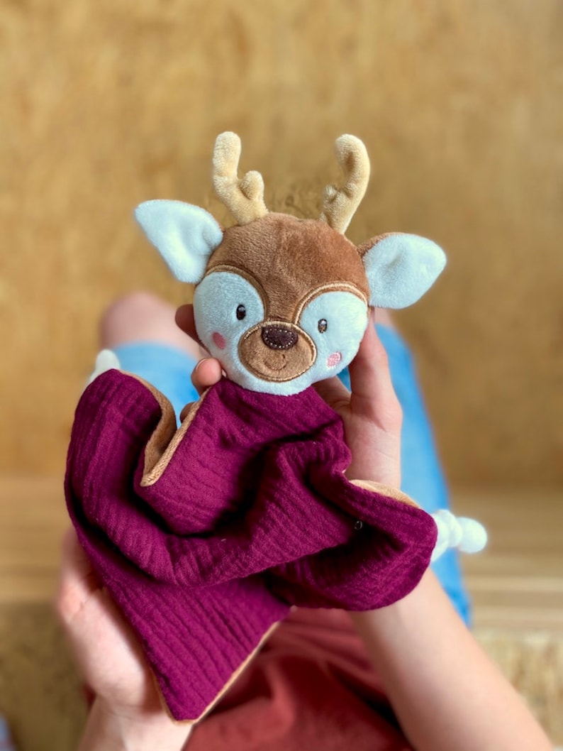 May include: A baby comforter in the shape of a deer. The deer's head is brown with beige antlers and white-lined ears. The comforter is a deep burgundy with a beige border. This soft toy is designed for infants and young children.