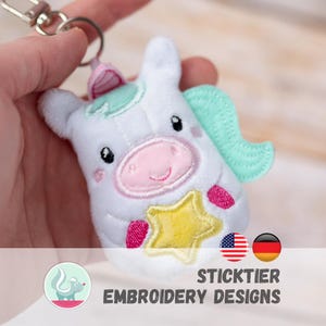 May include: A white plush unicorn keyring with a pink muzzle, a yellow star, and mint green accents. The keyring has a silver clasp and the text "STICKTIER EMBROIDERY DESIGNS" is visible at the bottom.