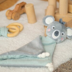 May include: A gray and mint green baby security blanket with a koala bear head. The blanket has a soft, plush texture and a cute koala face with embroidered details. A wooden toy car and a wooden ring teether are also visible.