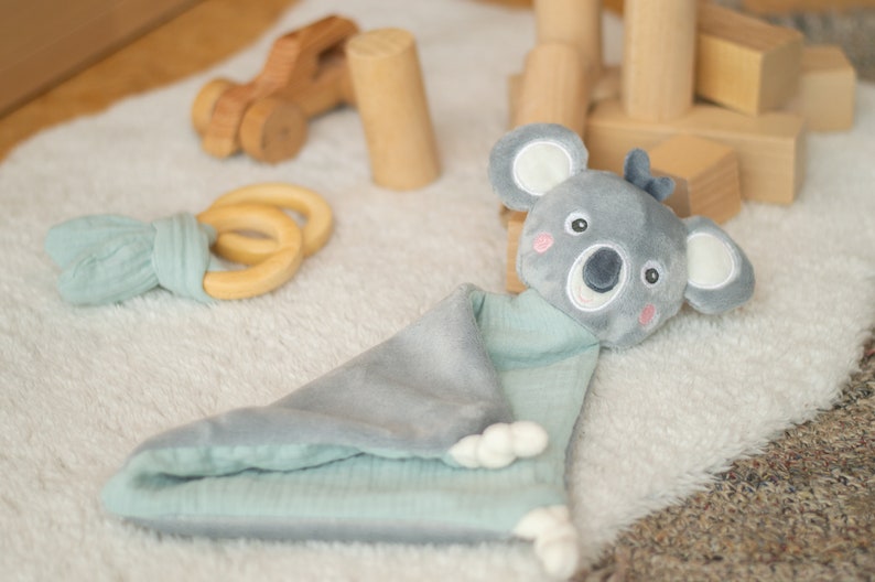 May include: A gray and mint green baby security blanket with a koala bear head. The blanket has a soft, plush texture and a cute koala face with embroidered details. A wooden toy car and a wooden ring teether are also visible.