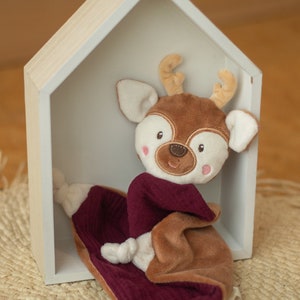 May include: A soft deer toy with a brown head, antlers, and body, wearing a claret scarf, and resting on a claret blanket. The toy is inside a white wooden house-shaped shelf. The background is a light brown.