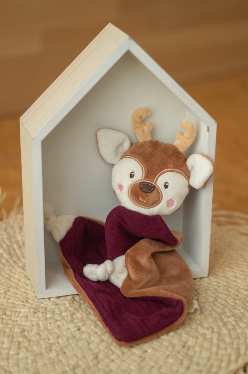 May include: A soft deer toy with a brown head, antlers, and body, wearing a claret scarf, and resting on a claret blanket. The toy is inside a white wooden house-shaped shelf. The background is a light brown.