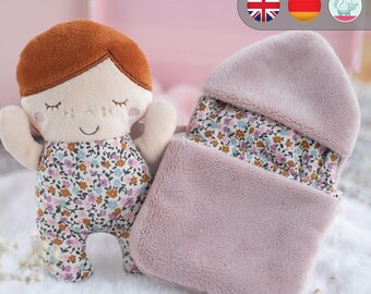 May include: A plush doll with brown hair and a floral dress, comes with a pink and white blanket with a hood. The doll is a girl with closed eyes and a smile. The blanket is made of soft, fuzzy material.