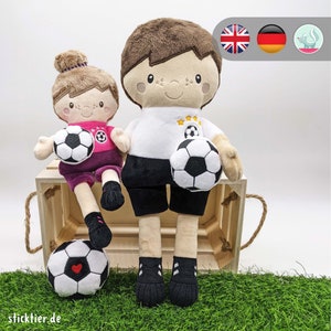 Può includere: Due giocatori di calcio di peluche, uno con una maglia bianca con pantaloncini neri e un pallone da calcio bianco e nero, l'altro con una maglia rosa con pantaloncini viola e un pallone da calcio bianco e nero. Entrambi sono seduti su una cassa di legno con un manico di corda.