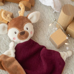 May include: A soft deer-shaped baby comforter with a brown head, cream-coloured accents, and a burgundy blanket. Wooden blocks and a wooden ring are scattered around the comforter on a soft, white surface. A string of lights with small white bulbs is also present.