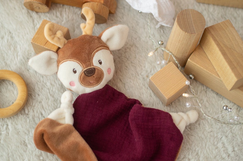 May include: A soft deer-shaped baby comforter with a brown head, cream-coloured accents, and a burgundy blanket. Wooden blocks and a wooden ring are scattered around the comforter on a soft, white surface. A string of lights with small white bulbs is also present.