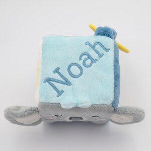 May include: A soft, plush baby block in shades of blue, gray, and yellow, with the name "Noah" embroidered in blue. The block features a koala face and ears, and a yellow propeller.