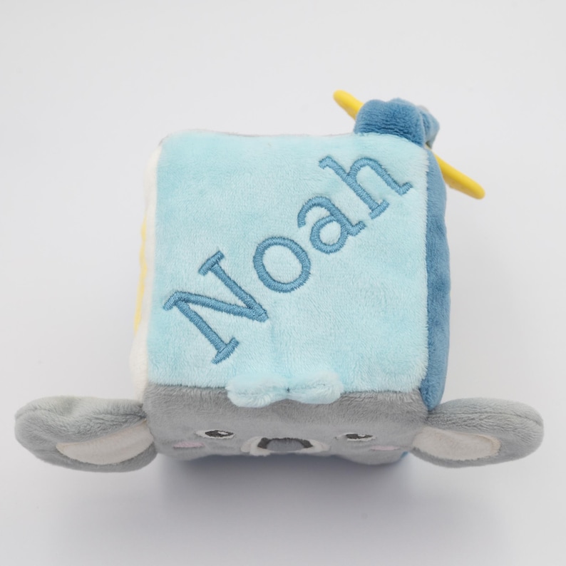May include: A soft, plush baby block in shades of blue, gray, and yellow, with the name "Noah" embroidered in blue. The block features a koala face and ears, and a yellow propeller.