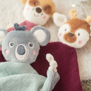 May include: A collection of soft, plush baby toys featuring animal faces. The toys include a gray koala, a brown deer, and a tan dog. Each toy has embroidered facial features and is set against a soft, textured background.