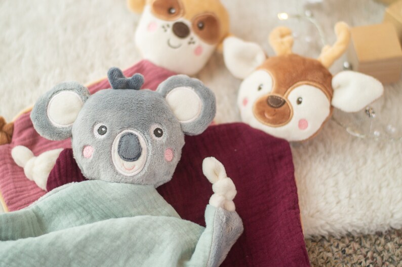 May include: A collection of soft, plush baby toys featuring animal faces. The toys include a gray koala, a brown deer, and a tan dog. Each toy has embroidered facial features and is set against a soft, textured background.