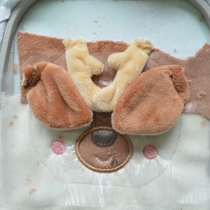 May include: An embroidery project in progress, depicting a bear's face. The design incorporates brown and beige fur elements for the ears and muzzle, with pink cheeks and a stitched brown nose and mouth. The fabric is held in an embroidery hoop.