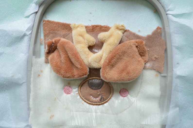 May include: An embroidery project in progress, depicting a bear's face. The design incorporates brown and beige fur elements for the ears and muzzle, with pink cheeks and a stitched brown nose and mouth. The fabric is held in an embroidery hoop.