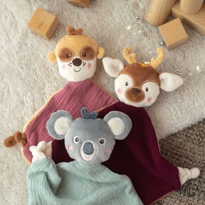 May include: Three plush baby comforters with animal faces: a raccoon, a deer, and a koala. The comforters have soft, textured blankets in shades of pink, teal, and burgundy. Wooden toy blocks and a wooden car are scattered around the comforters.