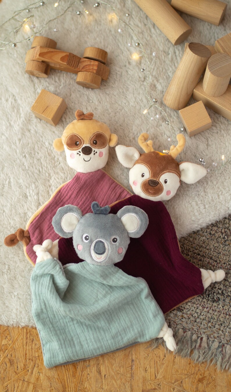 May include: Three plush baby comforters with animal faces: a raccoon, a deer, and a koala. The comforters have soft, textured blankets in shades of pink, teal, and burgundy. Wooden toy blocks and a wooden car are scattered around the comforters.