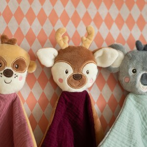 May include: Three plush animal security blankets: a tan dog, a brown deer, and a gray koala. Each has a soft, textured blanket in pink, burgundy, and light green. The animals have embroidered facial features and are set against a peach and white diamond pattern.