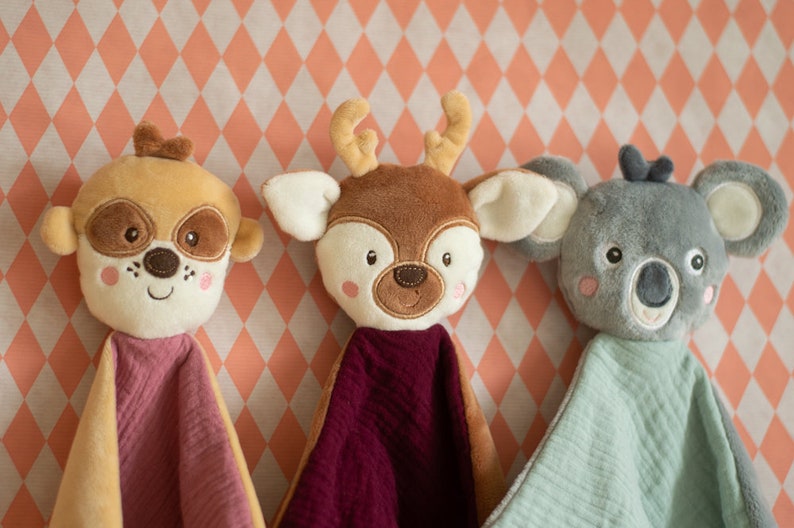 May include: Three plush animal security blankets: a tan dog, a brown deer, and a gray koala. Each has a soft, textured blanket in pink, burgundy, and light green. The animals have embroidered facial features and are set against a peach and white diamond pattern.