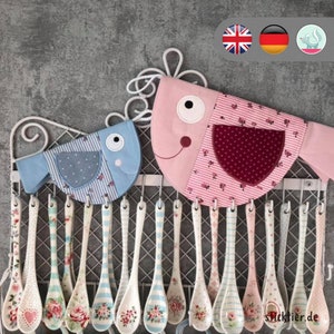 May include: A set of 20 white ceramic spoons with blue and white floral patterns hanging on a metal rack. Two fabric bird-shaped spoon holders are hanging on the rack. One holder is pink with white and red polka dots and the other is blue with white stripes.