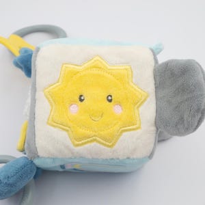 May include: A soft, plush baby toy cube with a smiling yellow sun design on one side. The cube features light blue, gray, and white panels, with attached plastic rings and a star-shaped accent. The toy is designed for infants and toddlers.