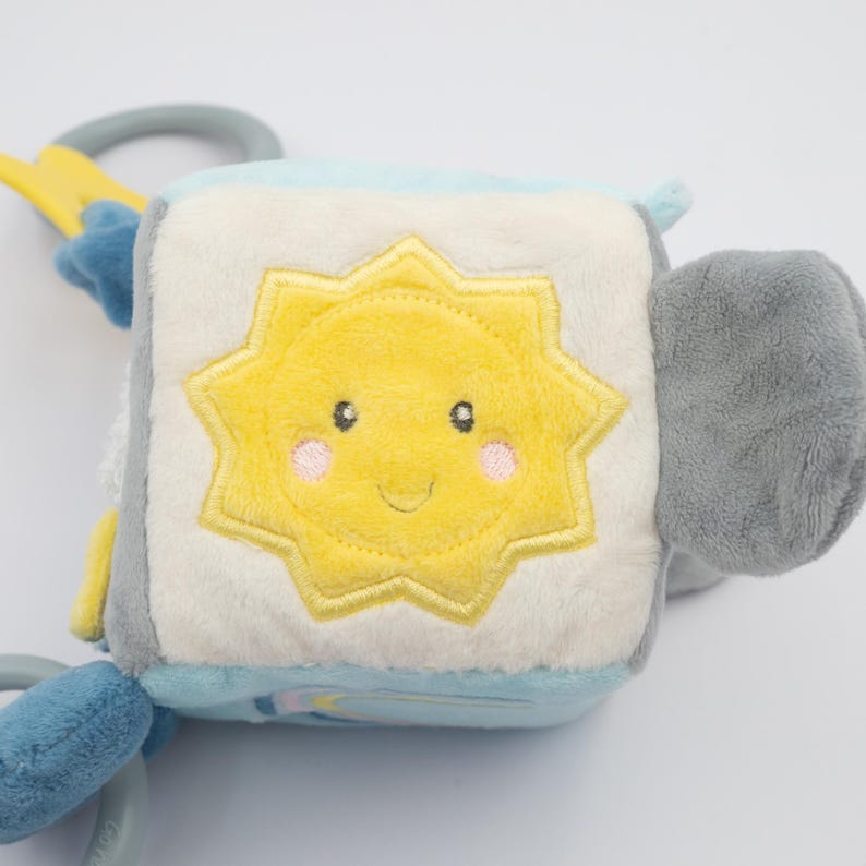 May include: A soft, plush baby toy cube with a smiling yellow sun design on one side. The cube features light blue, gray, and white panels, with attached plastic rings and a star-shaped accent. The toy is designed for infants and toddlers.