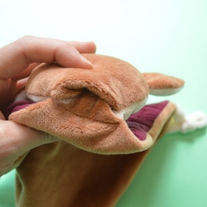 May include: A brown plush hand puppet with a light brown face, ears, and a burgundy interior. The puppet is held open, revealing the inside. The puppet's mouth is open, and the puppet is made of soft, plush material.