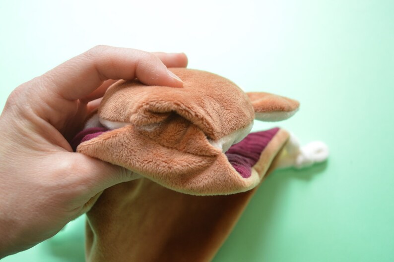 May include: A brown plush hand puppet with a light brown face, ears, and a burgundy interior. The puppet is held open, revealing the inside. The puppet's mouth is open, and the puppet is made of soft, plush material.