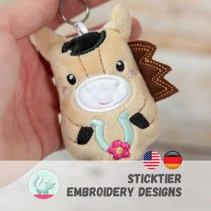 May include: A plush horse keychain in tan, brown, and black. The horse has a black hat, brown mane, and a horseshoe with a pink flower. The keychain is held by a hand. The text "STICKTIER EMBROIDERY DESIGNS" is at the bottom.