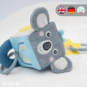 May include: A soft, grey koala-shaped baby toy with blue and yellow accents. The toy features embroidered details, a grey ring, and the text "STICKTIER.DE". The toy is designed with a soft, textured surface.