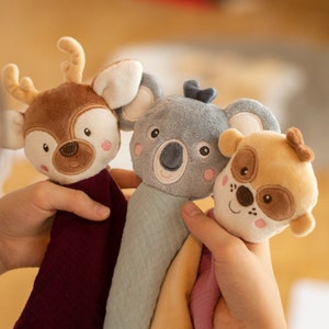 May include: Three plush animal toys: a deer, a koala, and a dog. The deer is brown with antlers, the koala is gray, and the dog is tan. Each has a soft, textured body in a coordinating color. The toys are held in a person's hands.