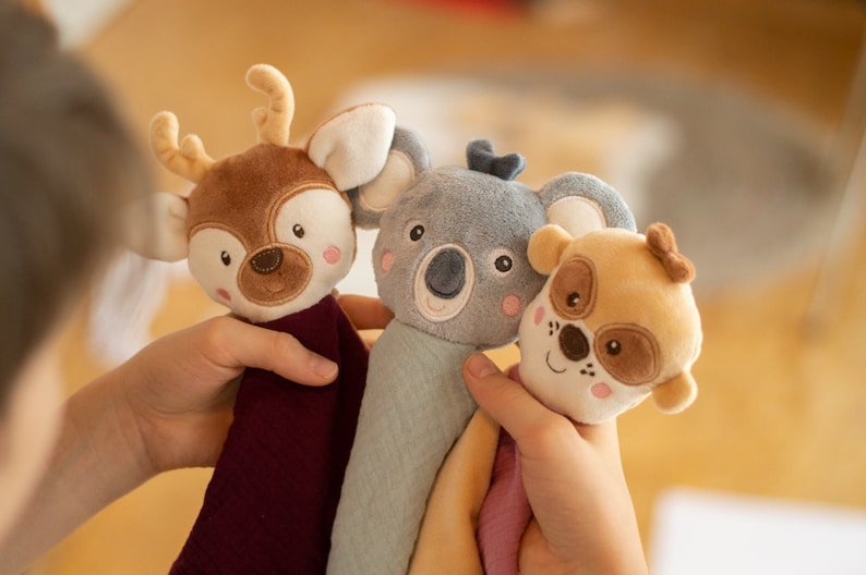May include: Three plush animal toys: a deer, a koala, and a dog. The deer is brown with antlers, the koala is gray, and the dog is tan. Each has a soft, textured body in a coordinating color. The toys are held in a person's hands.