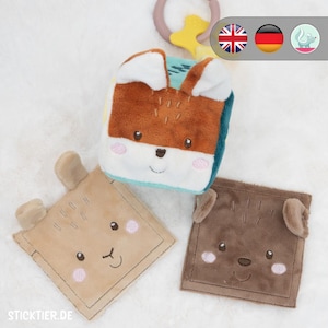 Puede incluir: Un conjunto de juguetes blandos para bebés con caras de animales. Los juguetes incluyen un cubo con forma de zorro y dos juguetes cuadrados con caras de oso. Los juguetes son en tonos de marrón, beige y azul, con detalles bordados y texturas suaves.