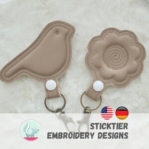 Könnte beinhalten: Zwei beige Leder-Schlüsselanhänger: einer in Vogelform und einer in Blumenform. Jeder Schlüsselanhänger hat einen Metallring und einen weißen Knopfverschluss. Sie liegen auf einer flauschigen weißen Oberfläche. Der Text "STICKTIER EMBROIDERY DESIGNS" befindet sich unten.